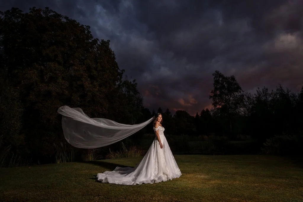 A woman in a wedding dress standing on grass during dusk with a long veil flowing behind her and dark stormy clouds in the sky.
