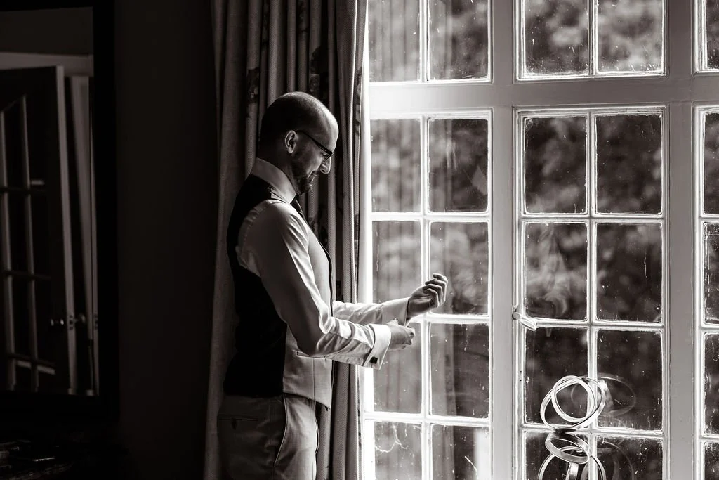 Black and white photo of a man in formal wear adjusting cuffs near a large window with multiple panes, sunlight streaming in, with a sculpture of rings on a table nearby.