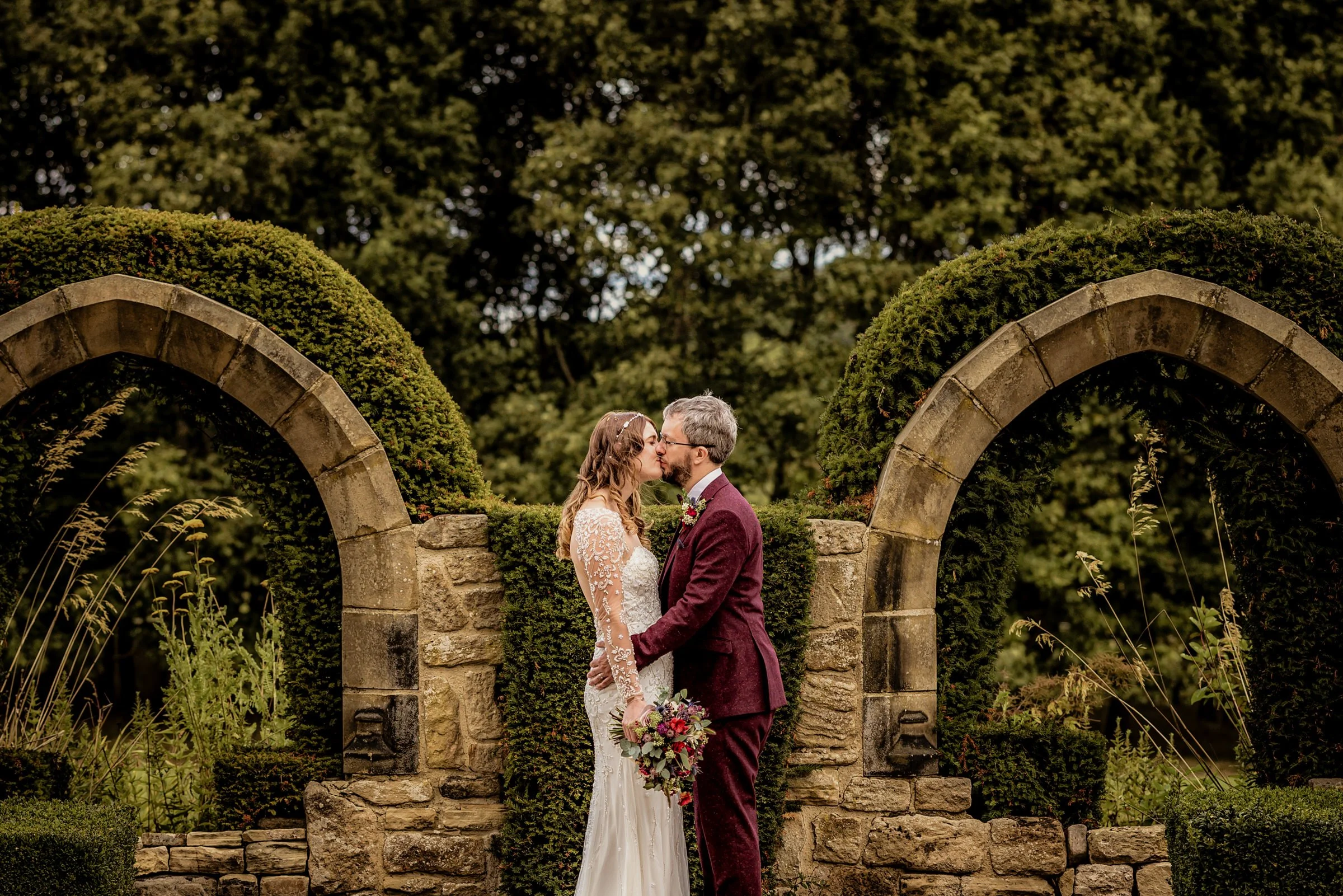 A bride and groom sharing a kiss in front of a stone archway surrounded by greenery in an outdoor wedding setting.