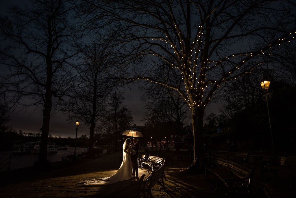 Couple with wedding attire standing under an umbrella at night, illuminated by a nearby streetlamp, with a tree decorated with string lights in the background.