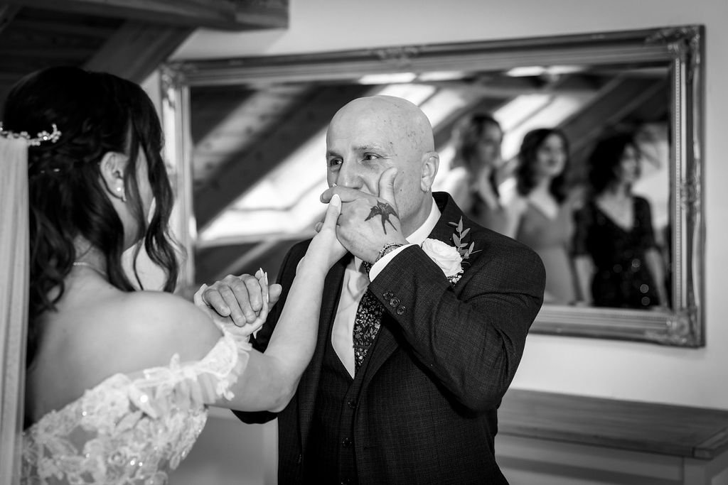 A bride and a groom during a wedding ceremony with the groom covering his mouth in emotion, a mirror reflecting three women in the background, one of whom is visible smiling.