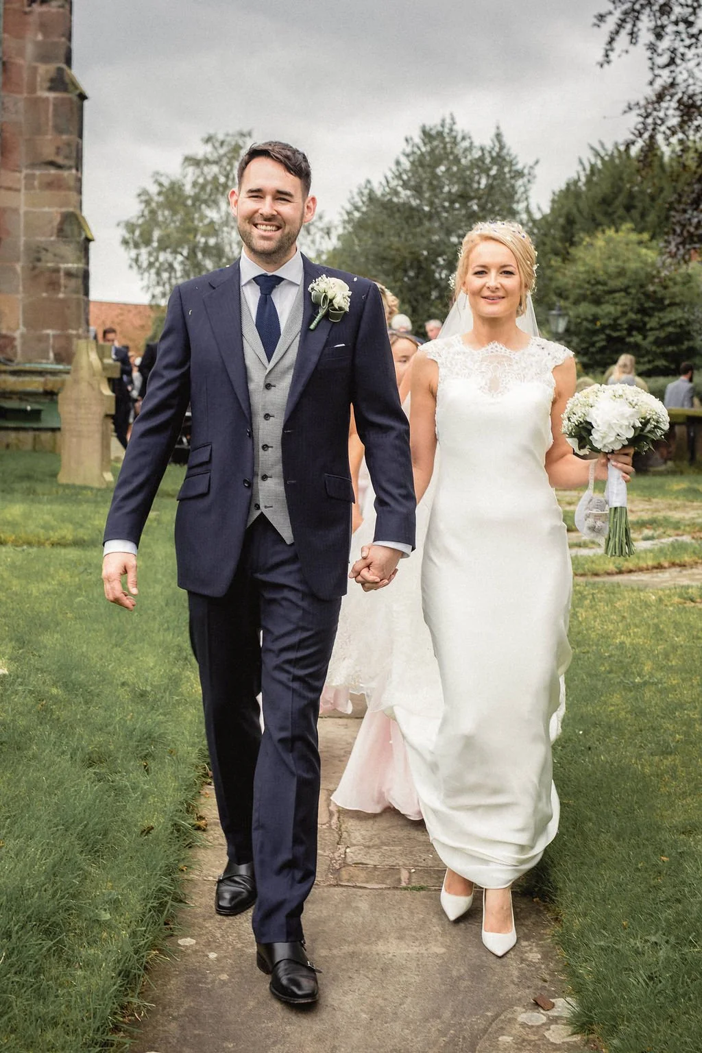 A bride and groom walking hand-in-hand outdoors on a cloudy day, with the bride holding a bouquet of white flowers. The groom is dressed in a dark blue suit, and the bride is in a white wedding gown with lace details.