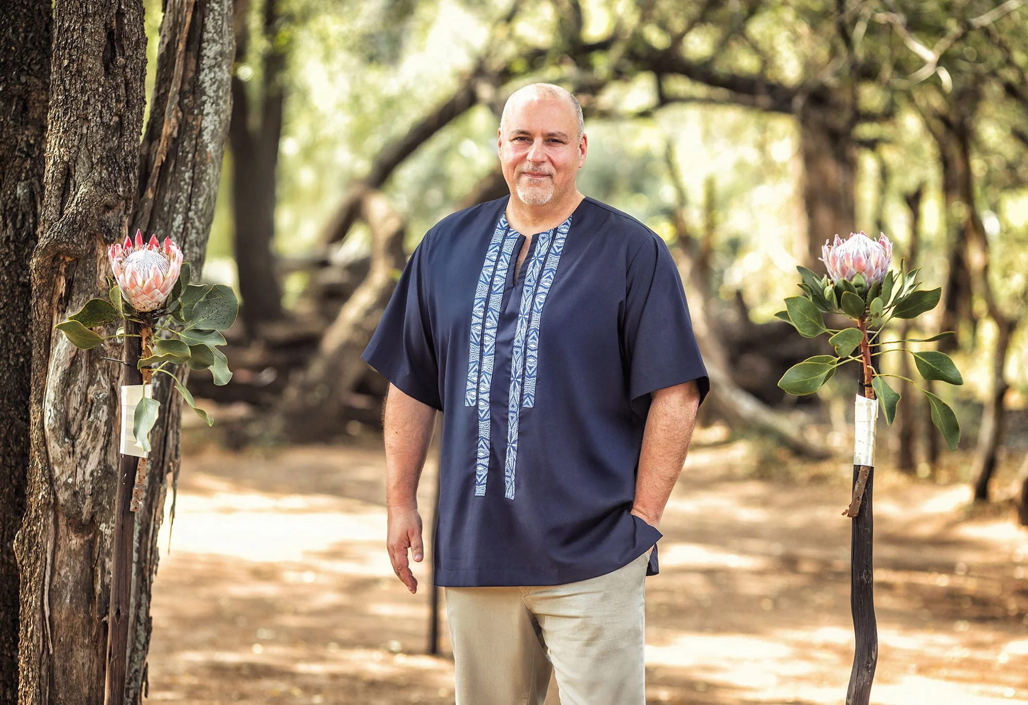 A middle-aged man with a goatee and a bald head standing outdoors in a wooded area, wearing a navy blue traditional shirt with blue patterns on the front, surrounded by trees and sunlight filtering through the leaves.