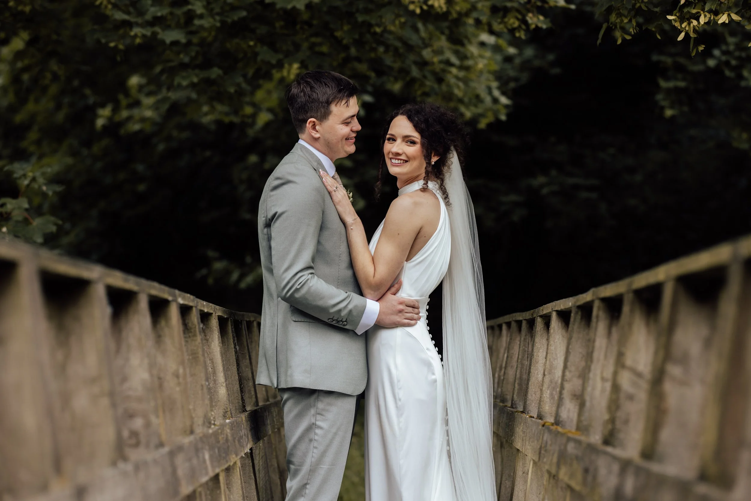 A smiling bride and groom standing close together on a wooden bridge surrounded by green trees during their wedding celebration.