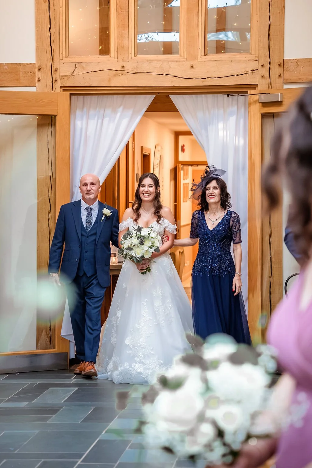 A bride in a white wedding gown holding a bouquet walking down the aisle with her parents in a wooden hall decorated for a wedding.
