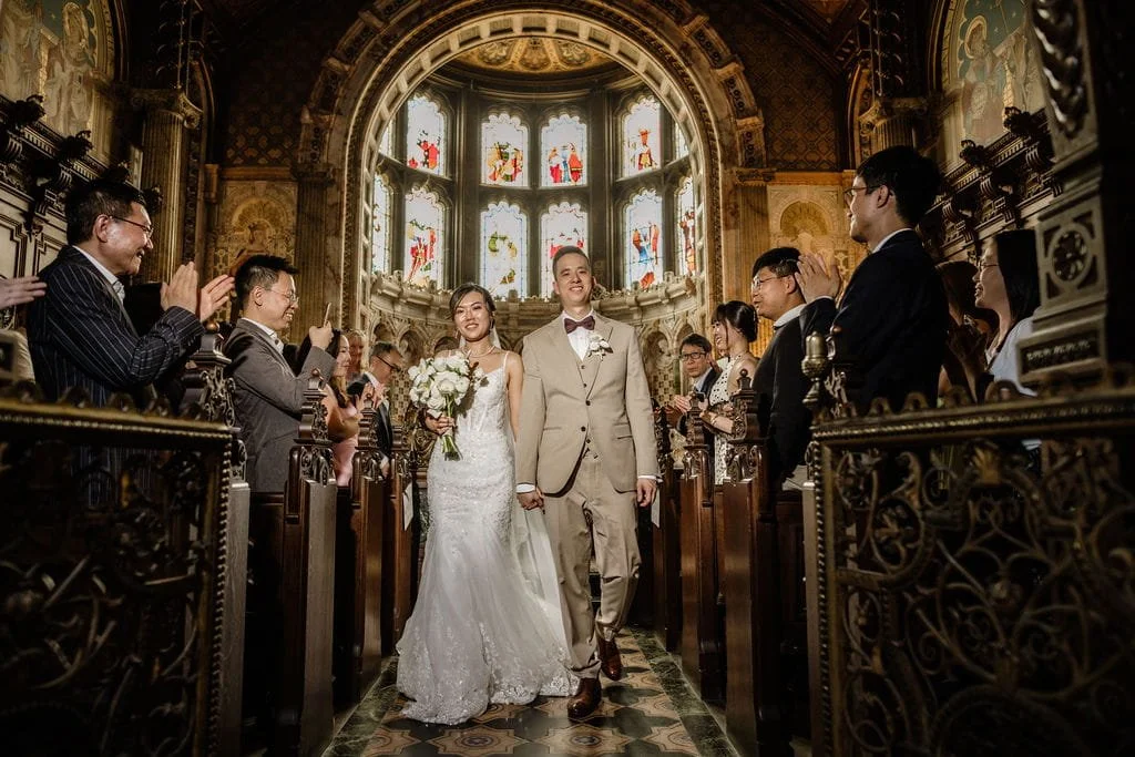 Bride and groom walking down the aisle in a decorated church filled with seated guests, stained glass windows are visible in the background.
