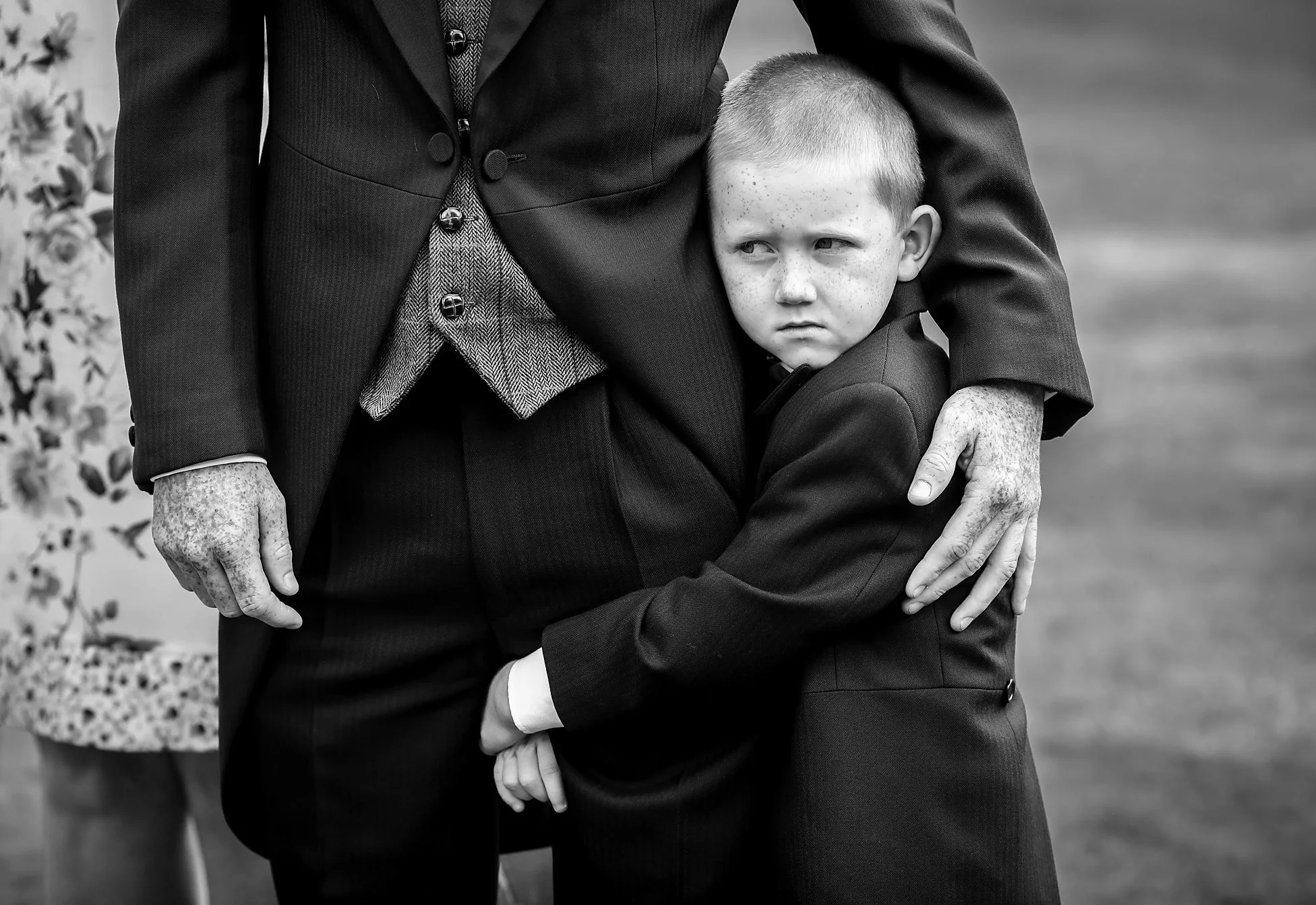 A young boy with freckles and a serious expression hugging an adult, possibly his father, standing outdoors. The boy's face is partially hidden as he rests his head against the adult's chest.
