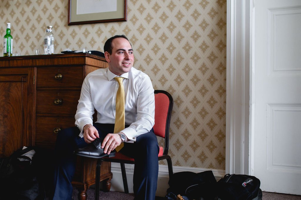A man in a white shirt and yellow tie sitting on a red and black chair in a room with patterned wallpaper. He is smiling, holding a cellphone, with a wooden cabinet behind him that has bottles and glasses on top. There are bags on the floor around hi