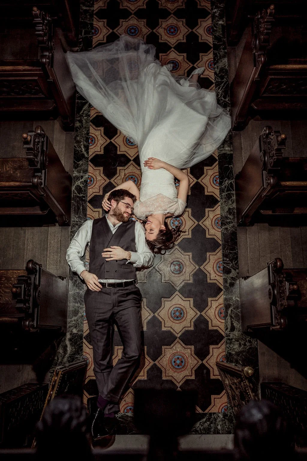 A bride and groom lying on a patterned church aisle floor, facing each other, with the bride in a white wedding gown and the groom in a gray vest and pants, surrounded by wooden church pews.