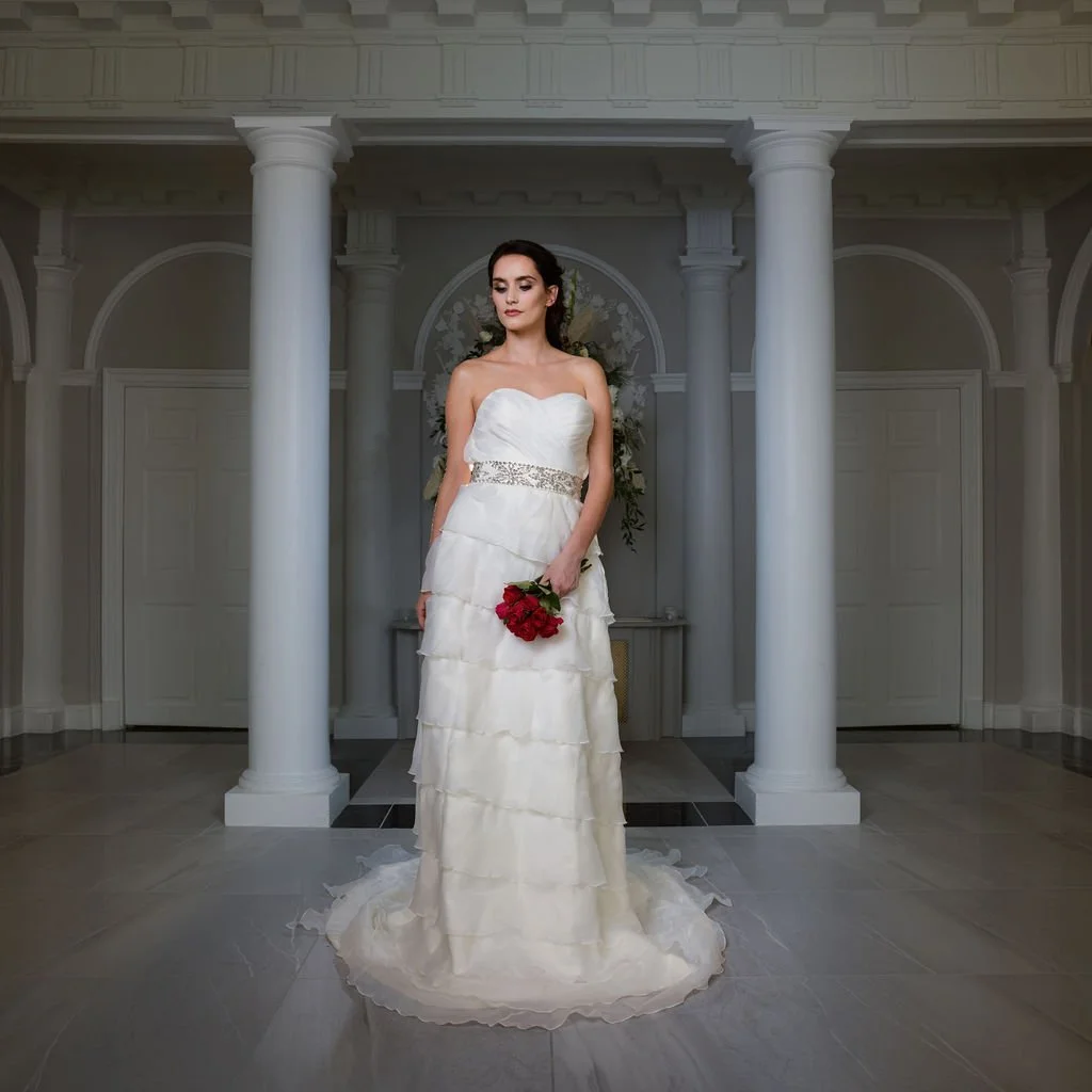 A woman in a white wedding dress holding a red bouquet in an elegant indoor setting with white columns and decorative architecture.