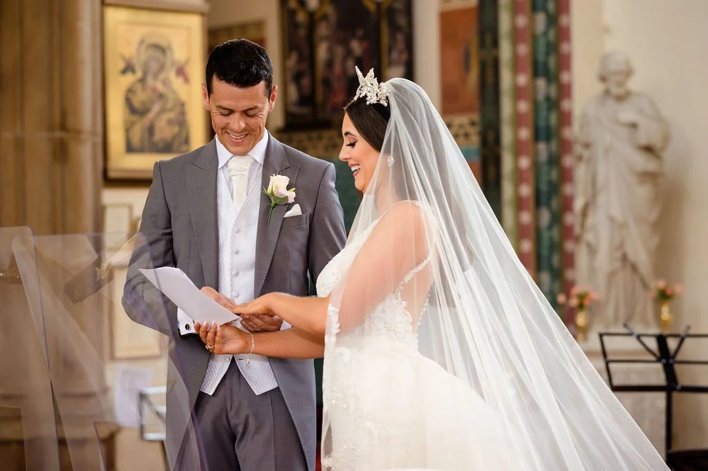 A bride and groom in a church during their wedding ceremony, exchanging vows or rings, with the bride smiling and wearing a veil and tiara, and the groom smiling in a gray suit.