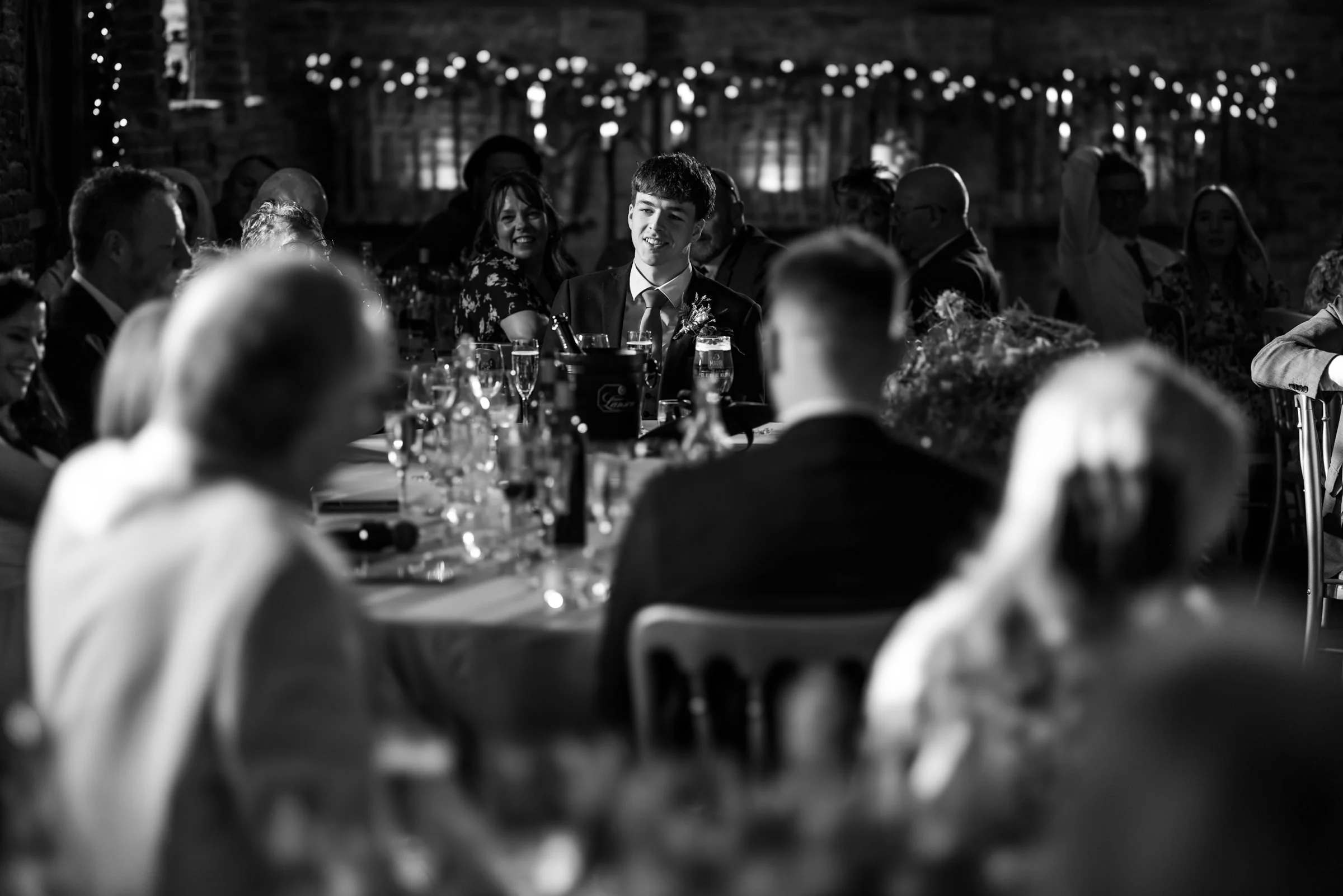 A black and white photo of a young man in a suit and tie at a formal event, sitting at a table with other guests. The setting appears to be a banquet or wedding reception with string lights in the background and glasses and bottles on the table.