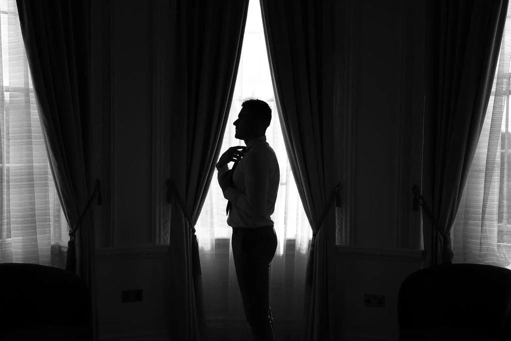 Silhouette of a man adjusting his tie in front of curtains and windows.