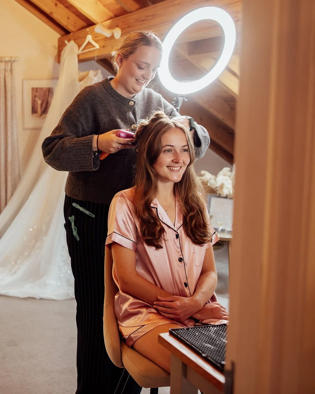 A woman getting her hair styled in a cozy, well-lit room with wooden walls and ceiling, wearing pink pajamas and smiling as a hairstylist, standing behind her, uses a curling iron. A ring light provides additional lighting, and a window with curtains