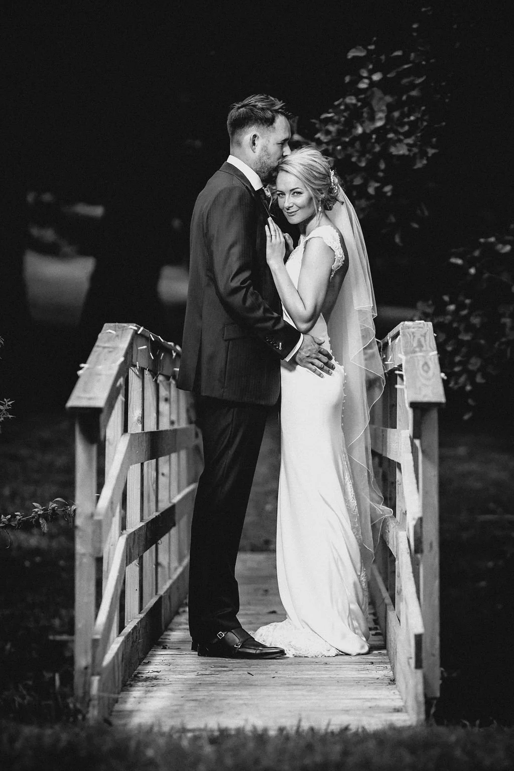 Black and white photo of a bride and groom standing on a small wooden bridge, embracing and gazing at each other, with a dark background and foliage.