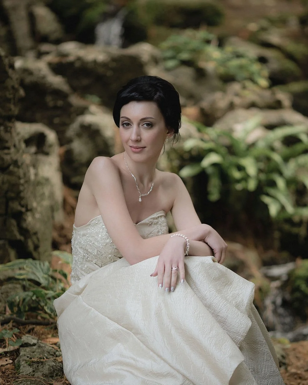 A woman in a strapless wedding dress sitting outdoors in a natural setting with rocks and greenery in the background.