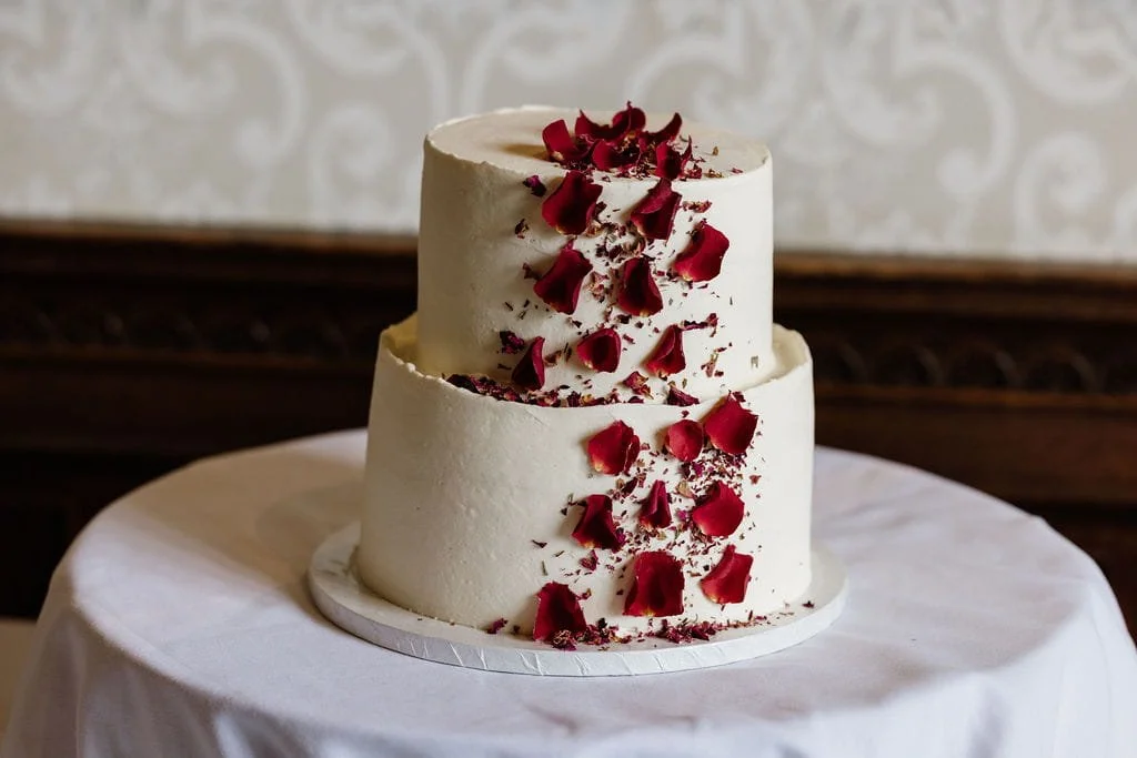 Two-tiered white wedding cake decorated with red rose petals on top and along the sides, placed on a white tablecloth.