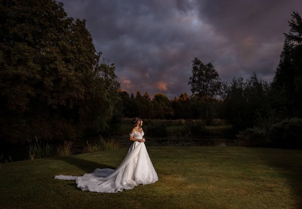 A woman in a white wedding dress standing on a grassy area near a pond at dusk, surrounded by trees and a dark cloudy sky.