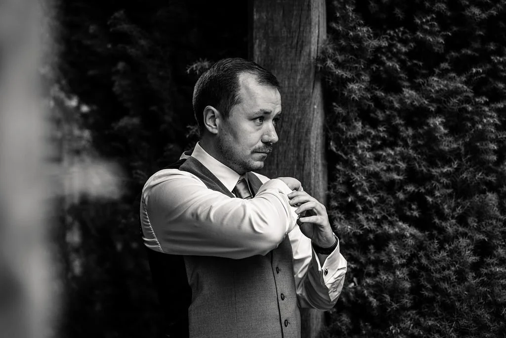 A man adjusting his sleeve in an outdoor setting with trees and a wooden post in the background.