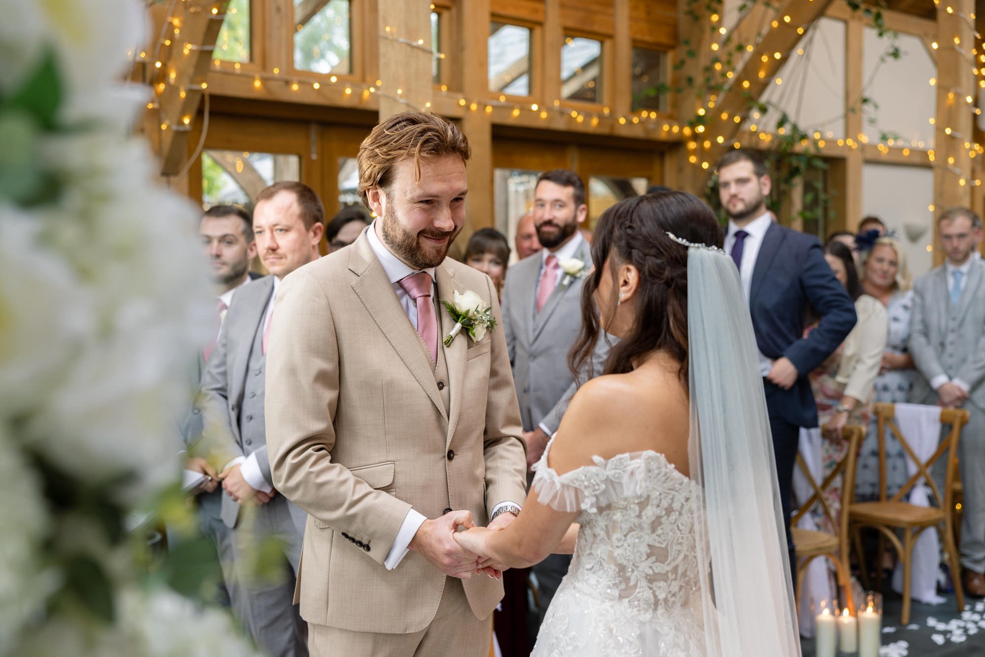 A bride and groom holding hands during their wedding ceremony, surrounded by guests in a decorated indoor venue with wooden accents and string lights.