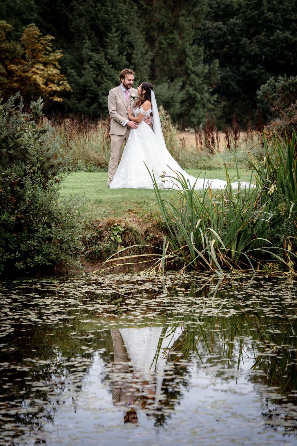 A newlywed couple stands on a grassy area beside a pond with floating leaves, surrounded by lush greenery and trees, during daytime. The bride wears a white wedding gown and veil, and the groom is in a light beige suit with a pink tie. They are looki