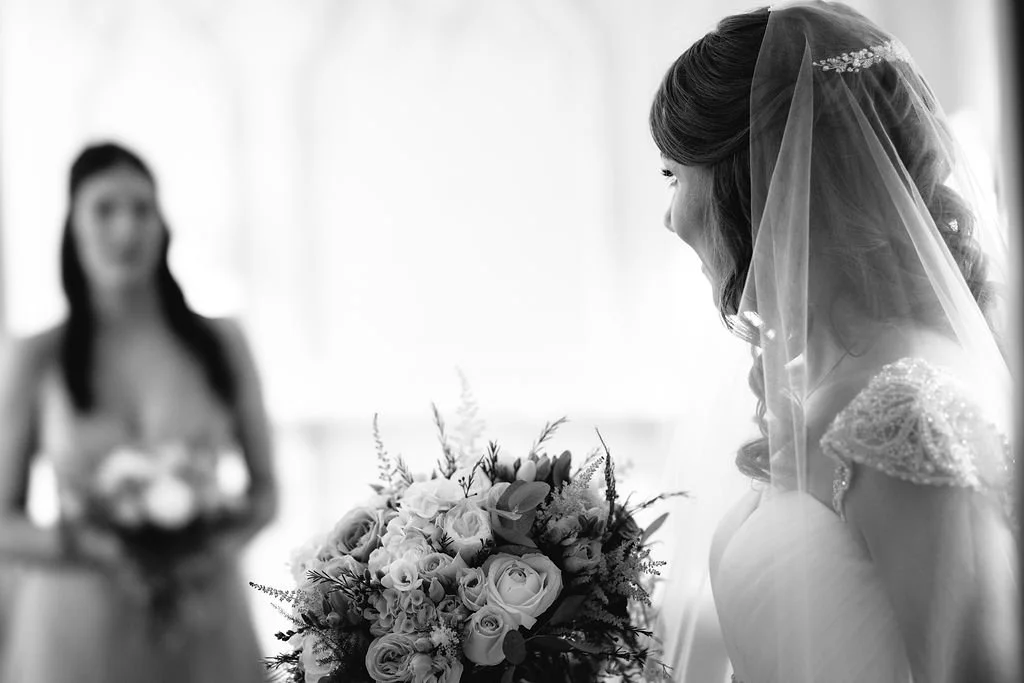 A bride with a veil and a wedding gown looking at a large bouquet of flowers. An out-of-focus woman in a dress is in the background holding a smaller bouquet.