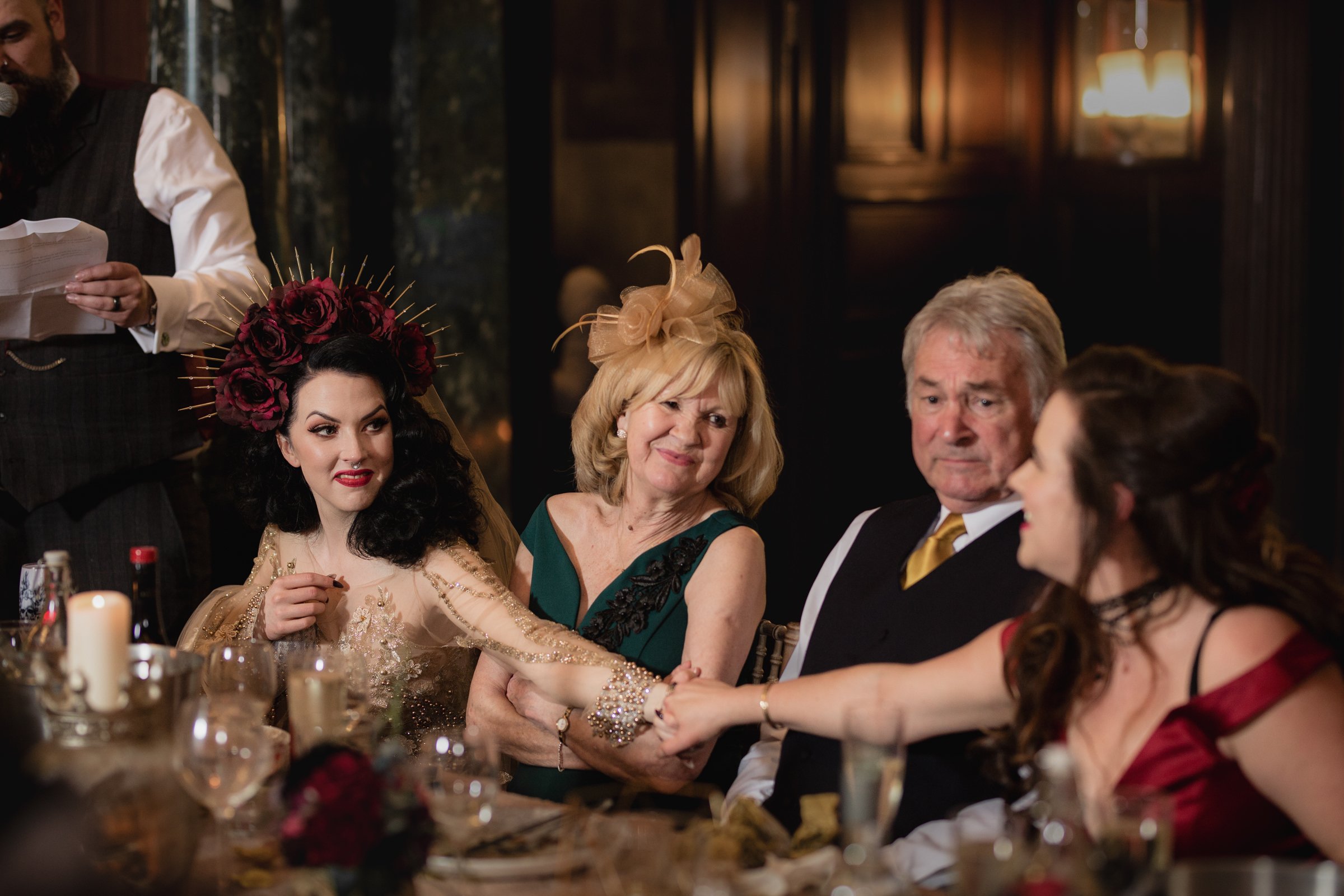 A group of five people sitting at a fancy dinner table during a celebration. The woman on the left wears a floral crown with dark red roses and a beige dress with gold embroidery. The woman next to her has blonde hair, wearing a green dress with blac
