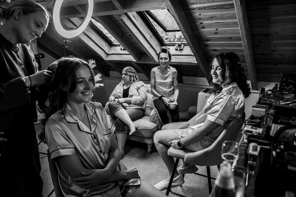 Women getting ready in a room with a skylight, one is having her hair styled by a makeup artist, others seated and smiling.