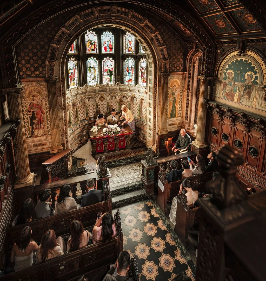 A wedding ceremony taking place inside an ornate church with stained glass windows, religious iconography, and a congregation seated in wooden pews.