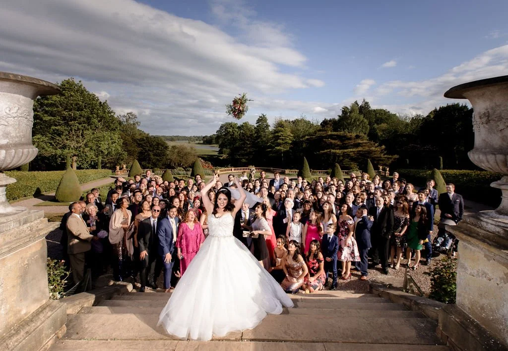 A bride in a white wedding dress standing on the steps of an outdoor wedding venue, surrounded by numerous wedding guests, with a scenic landscaped background and a blue sky with clouds.