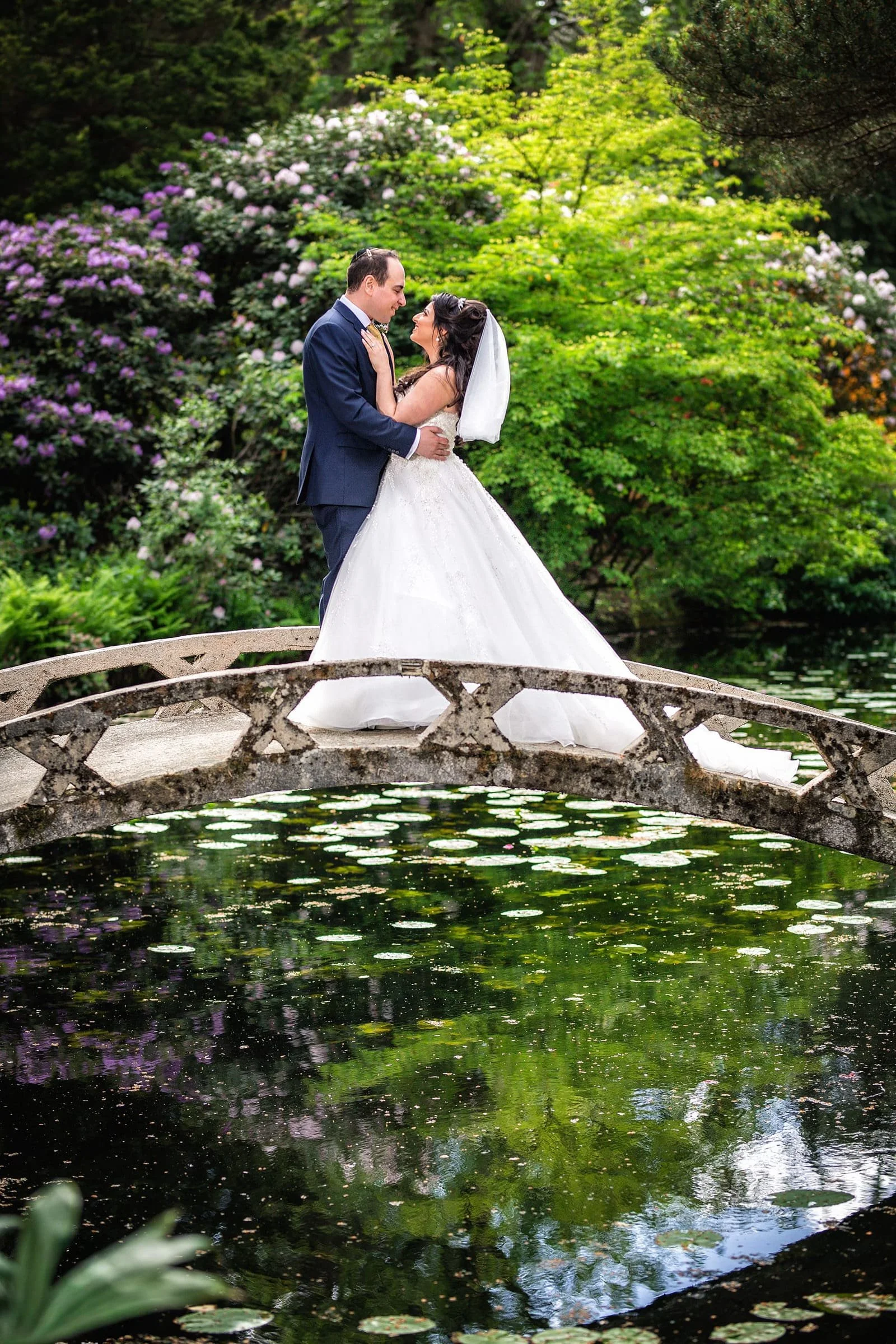 Couple in wedding attire standing on a small stone bridge over a pond, surrounded by lush green trees and flowering bushes.