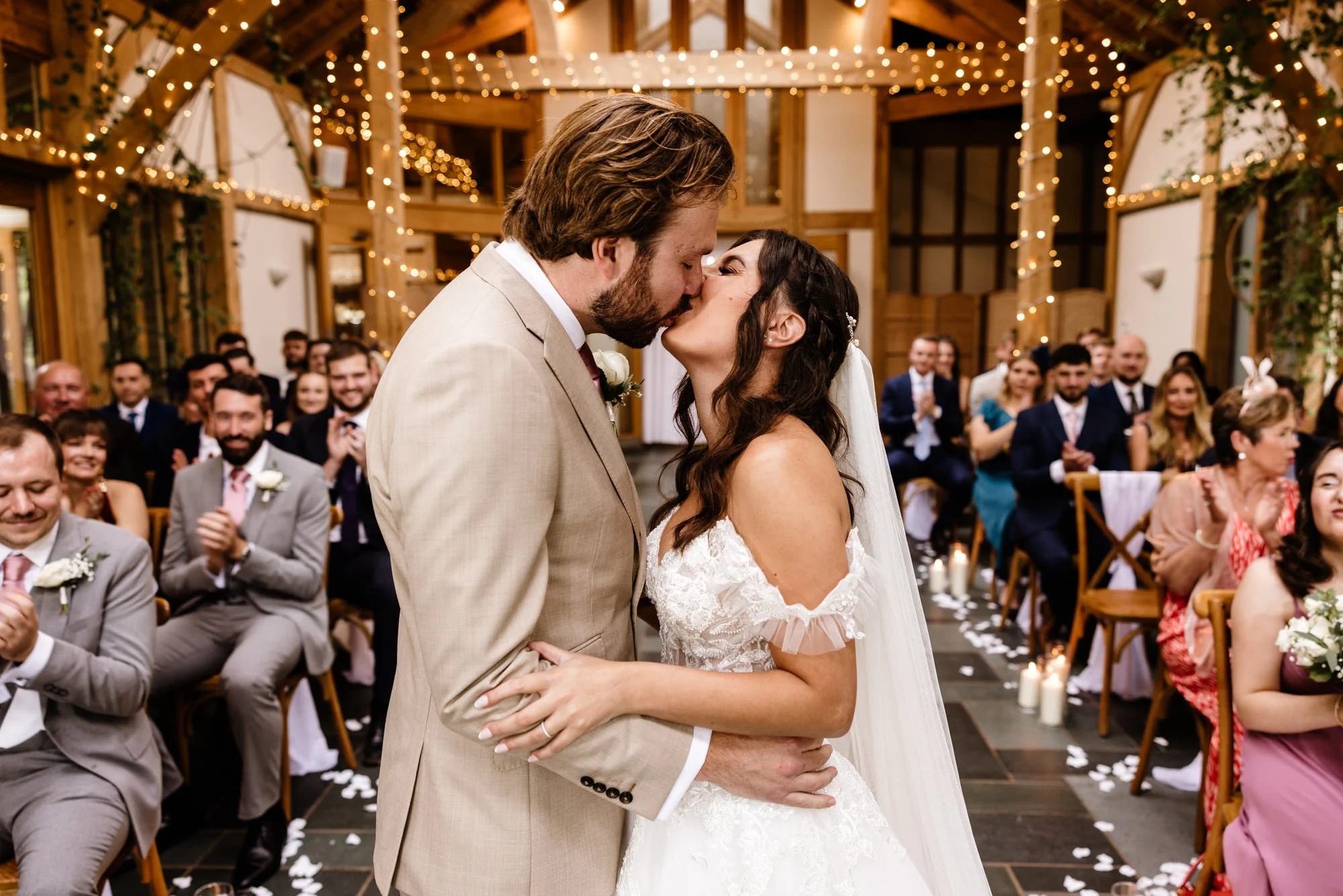 A newlywed couple shares a kiss during their wedding ceremony, surrounded by friends and family seated in a decorated wooden venue with string lights and candles.