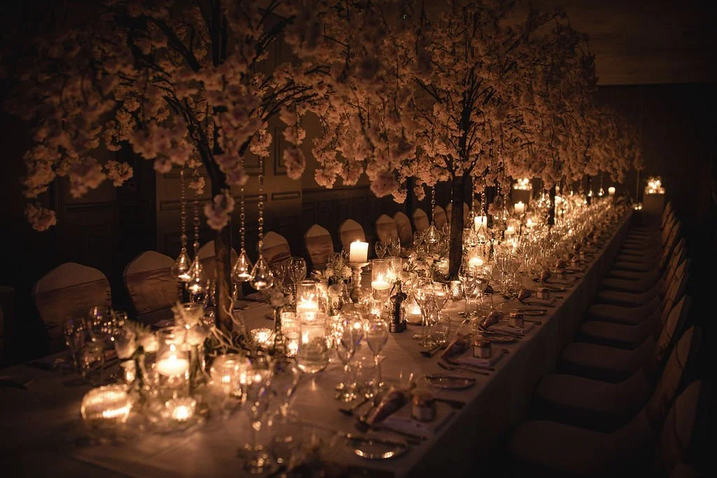 Long dinner table decorated with white flowers, candles, and glassware, set for a formal event in a dimly lit room.