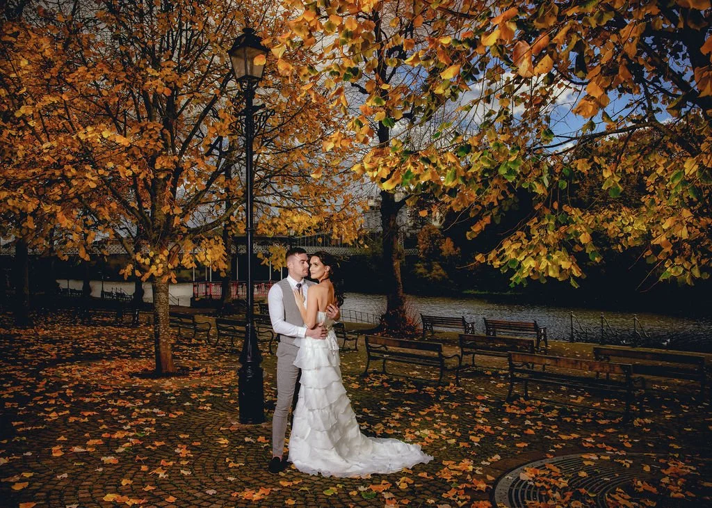 A couple in wedding attire standing close together in a park during autumn, surrounded by trees with orange and yellow leaves, near a river with benches along the path.