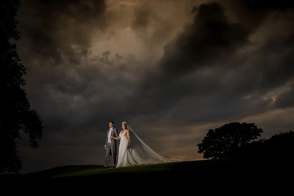 A bride and groom standing outdoors at night, under a dark, cloudy sky. The bride is in a wedding dress with a long train and veil, holding the bridegroom's arm. The groom is in a suit, standing beside her.