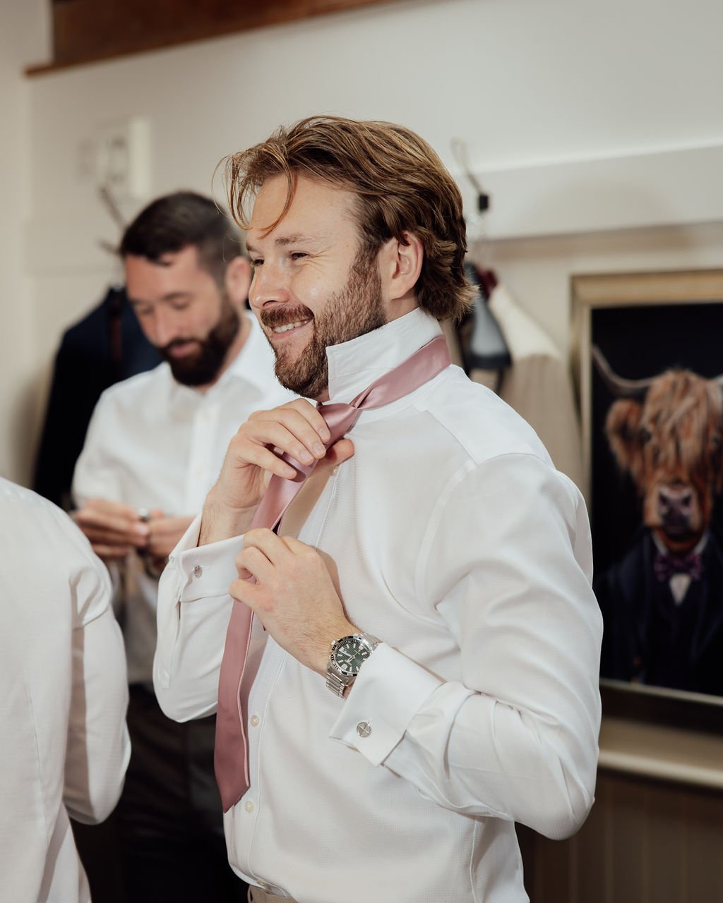 A man in a white dress shirt and pink tie adjusting his tie, smiling, with another man in white shirt and a person in a tiger mask in the background.