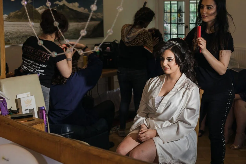 A bride sitting with her hands on her lap while a woman styles her hair and applies makeup in a room with multiple people preparing for a wedding.
