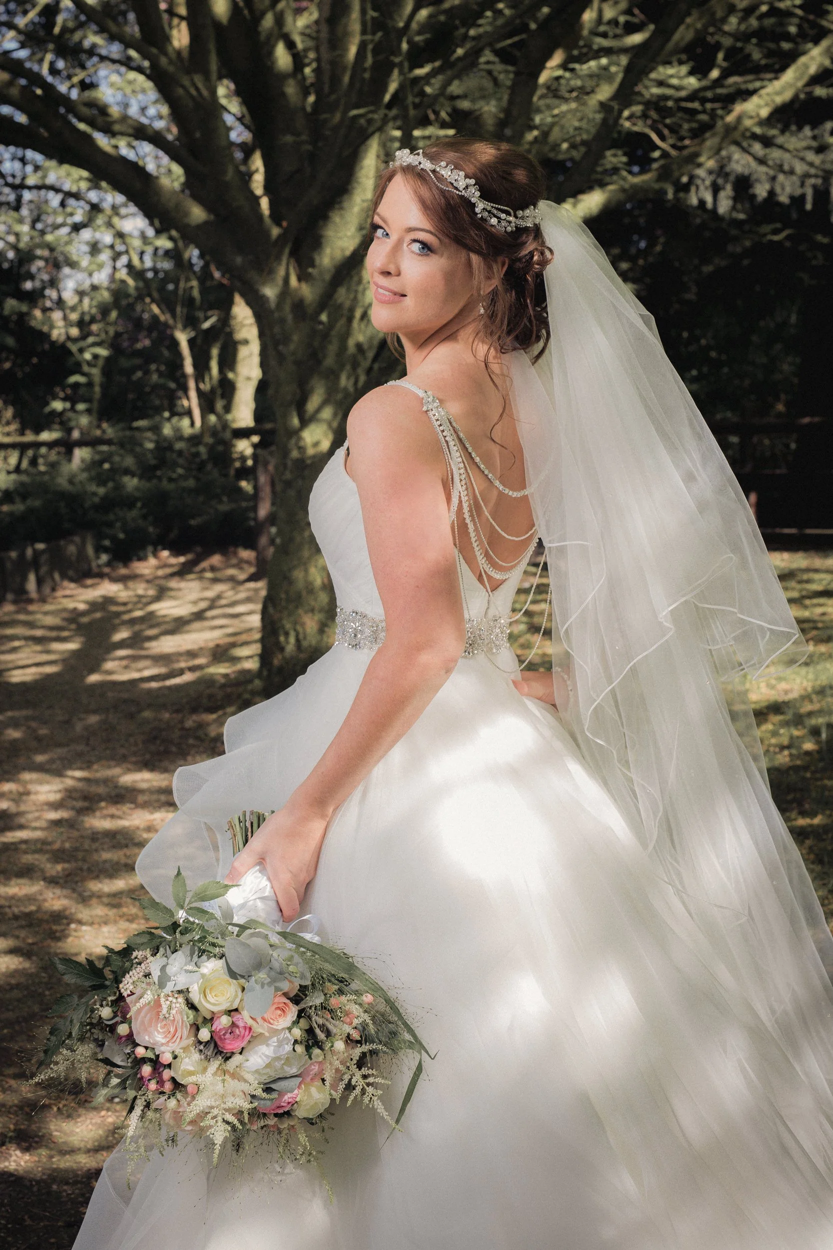 Brunette bride in a white wedding dress with beaded straps and a long veil, holding a bouquet of white, pink, and cream roses with greenery, standing outdoors under a large tree with textured bark and lush branches.