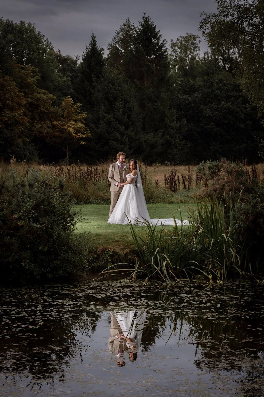 A bride and groom holding hands in wedding attire outdoors near a pond with reflections of trees and their couple.