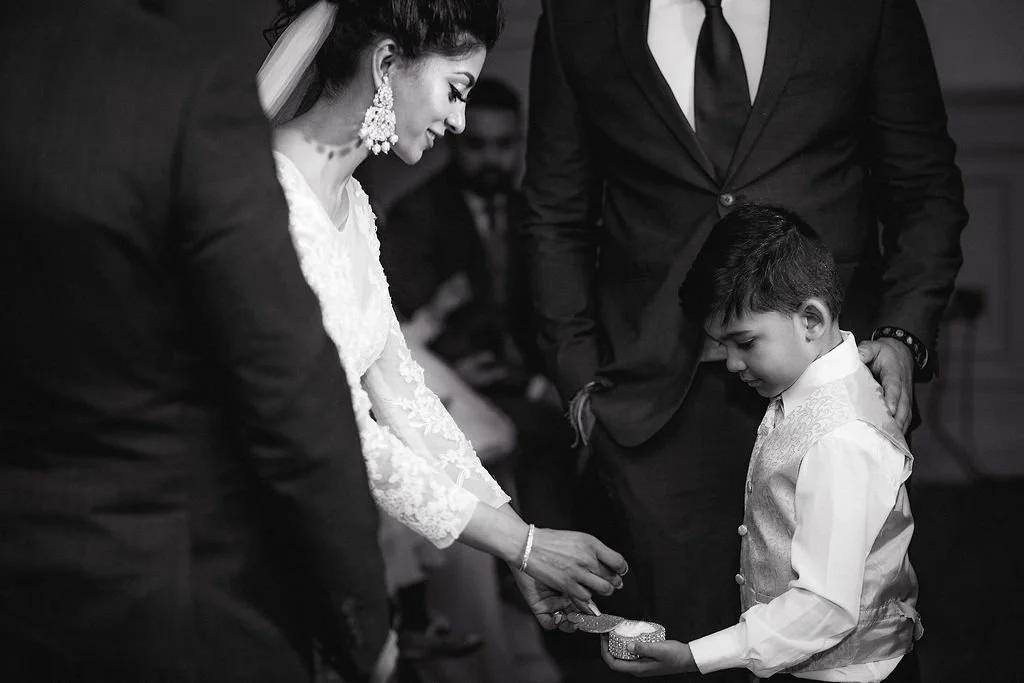 A woman in a wedding dress is holding a wedding ring, and a young boy in a vest is holding a jewelry box, during a wedding ceremony, with men in suits nearby.