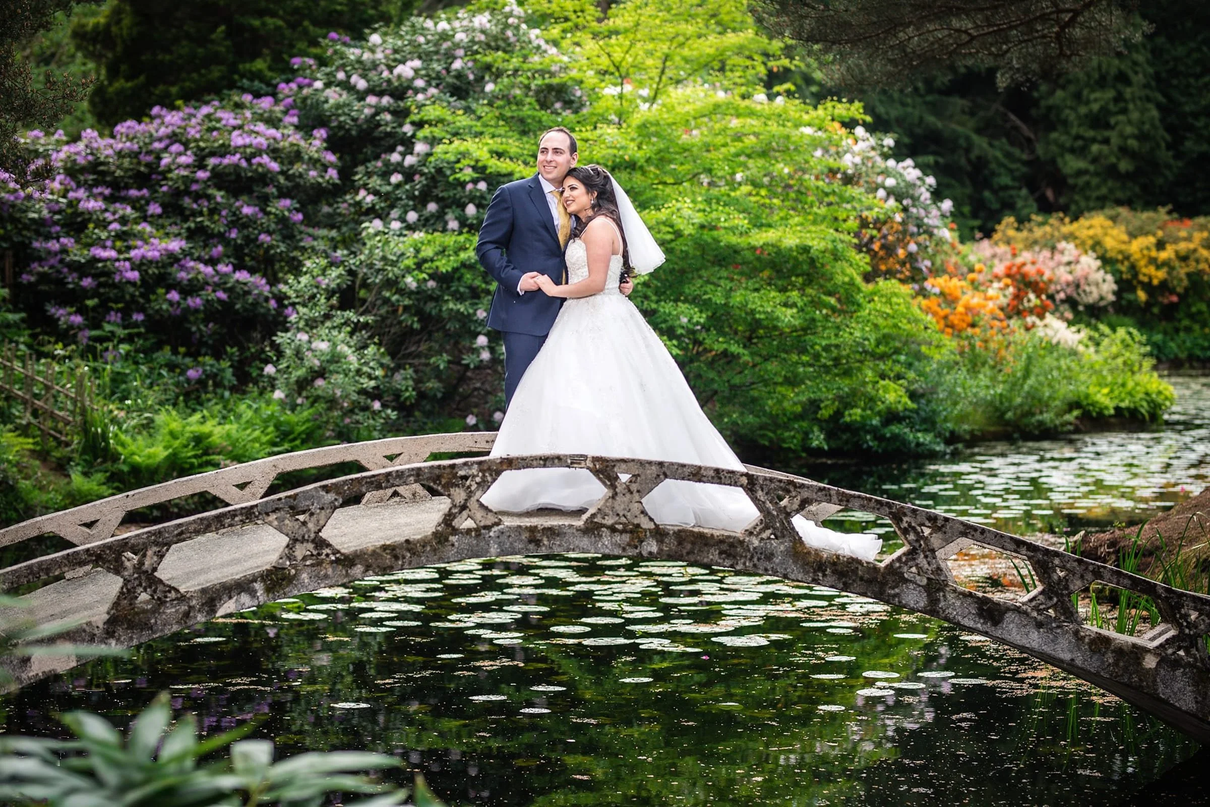 A bride and groom standing close together on a small stone bridge over a pond in a lush garden, surrounded by green trees and colorful blooming flowers.
