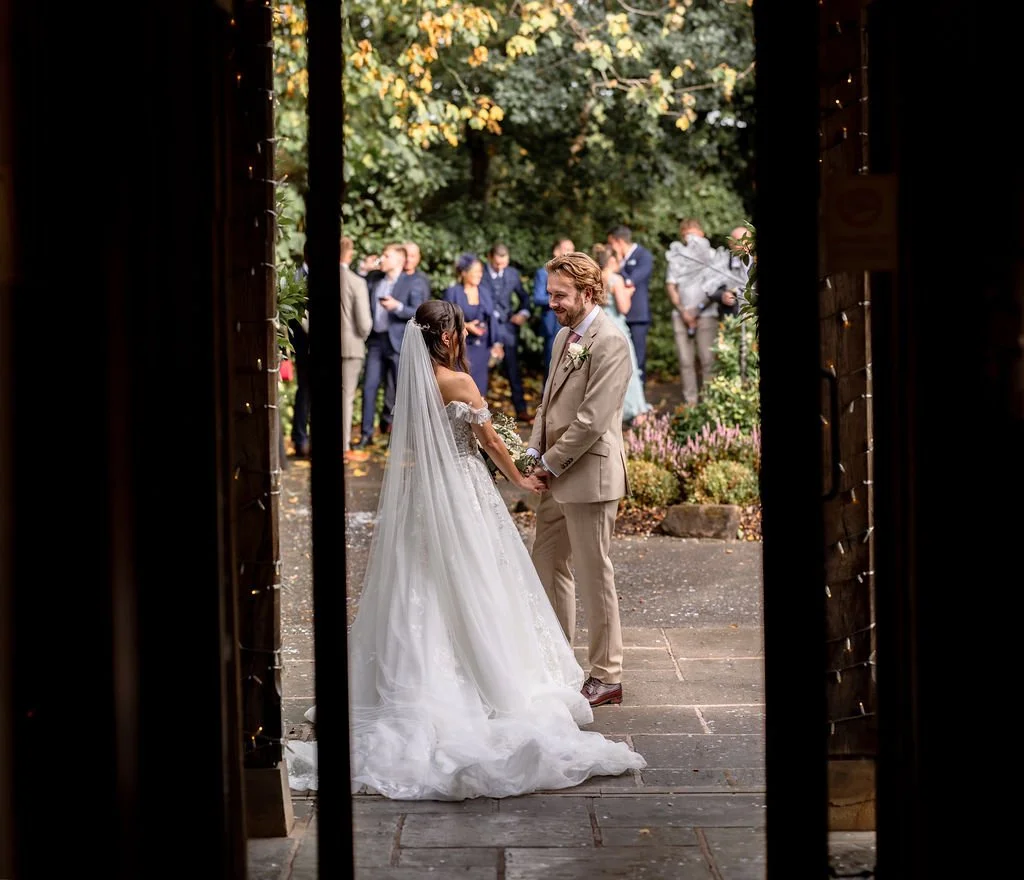 Bride and groom holding hands during their outdoor wedding ceremony, with guests in the background and trees surrounding the area.