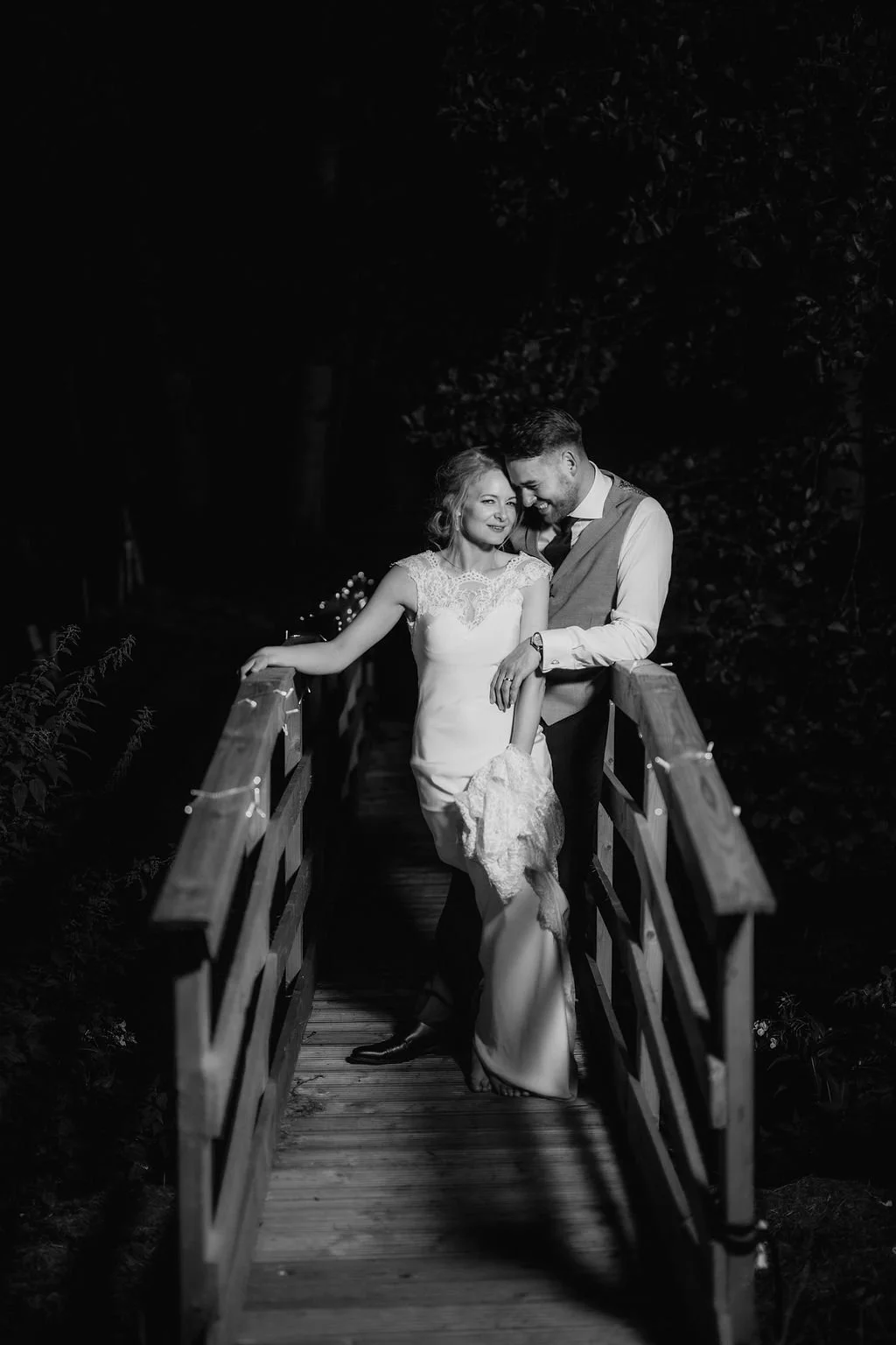 Black and white photo of a bride and groom standing together on a small wooden bridge at night, embracing and smiling.