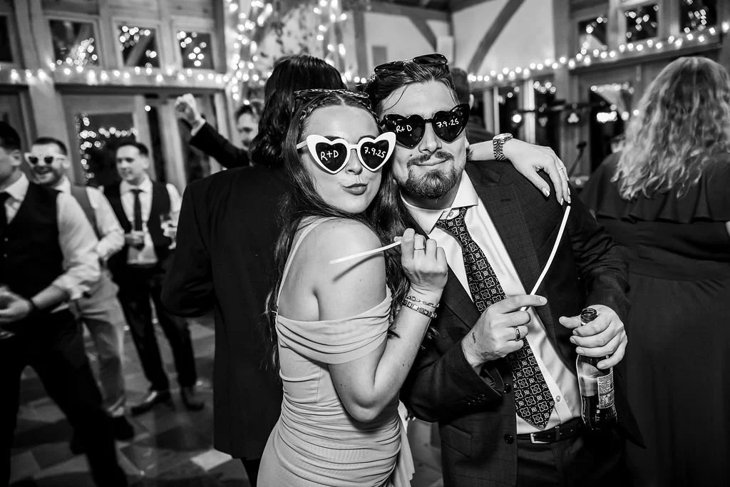 People celebrating at a party, with two central figures wearing heart-shaped sunglasses and holding drinks, smiling and embracing; festive decorations and other guests in the background.