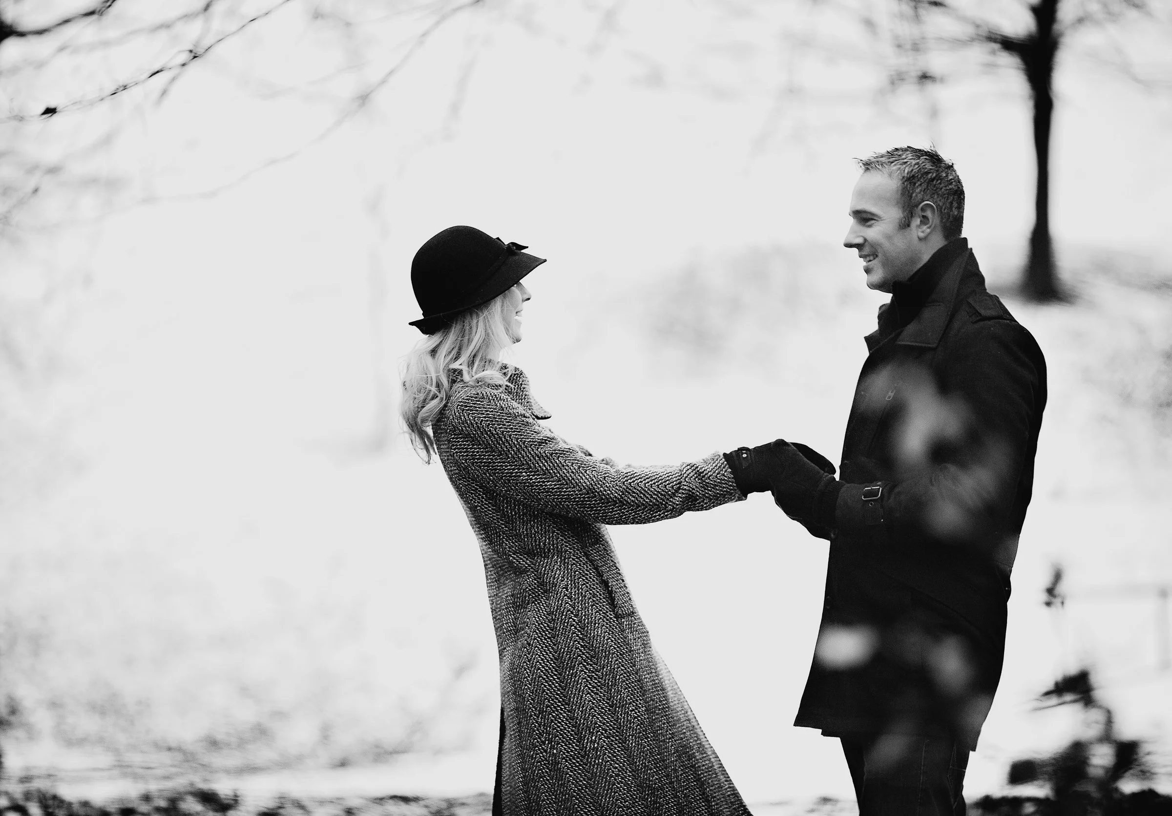 A couple holding hands and smiling at each other outdoors on a winter day, with trees and snow in the background, in black and white.