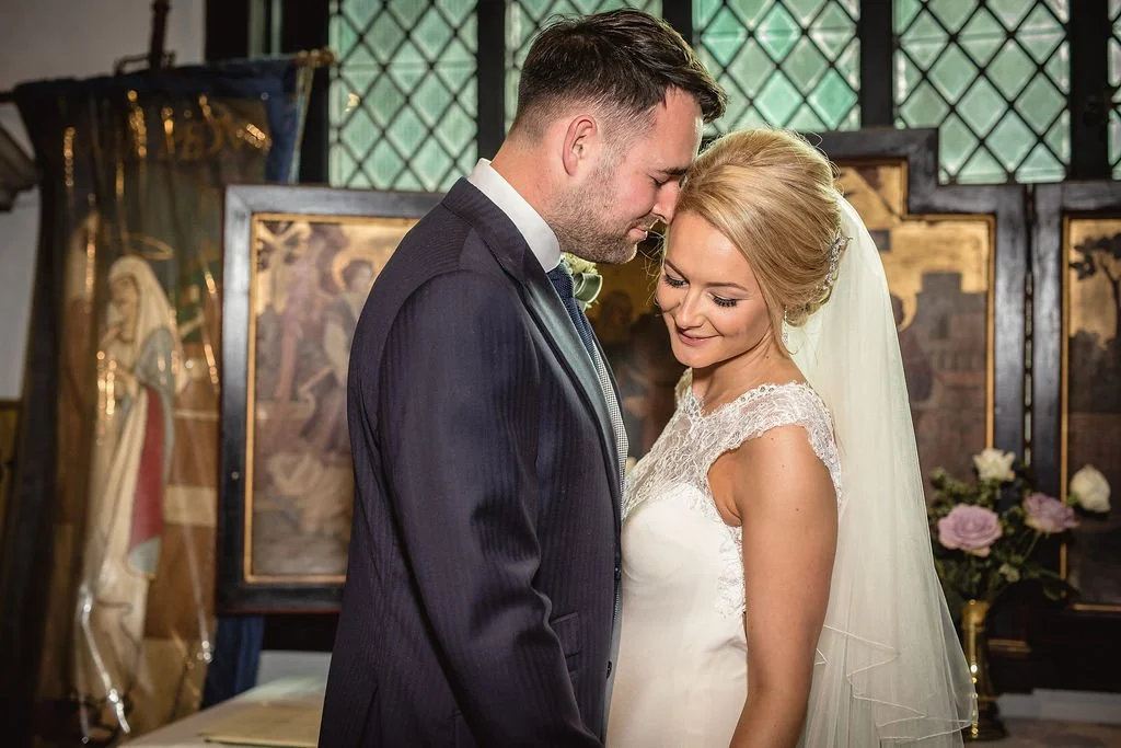 A bride and groom are standing close with their foreheads touching in a church, with religious artwork and flowers in the background.