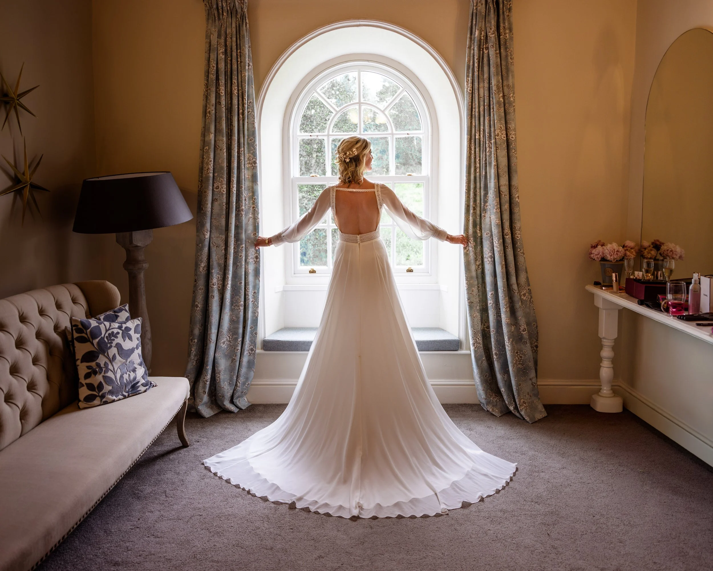 Bride in a flowing white wedding dress standing by a window, looking outside, with floral curtains in a warmly lit room.