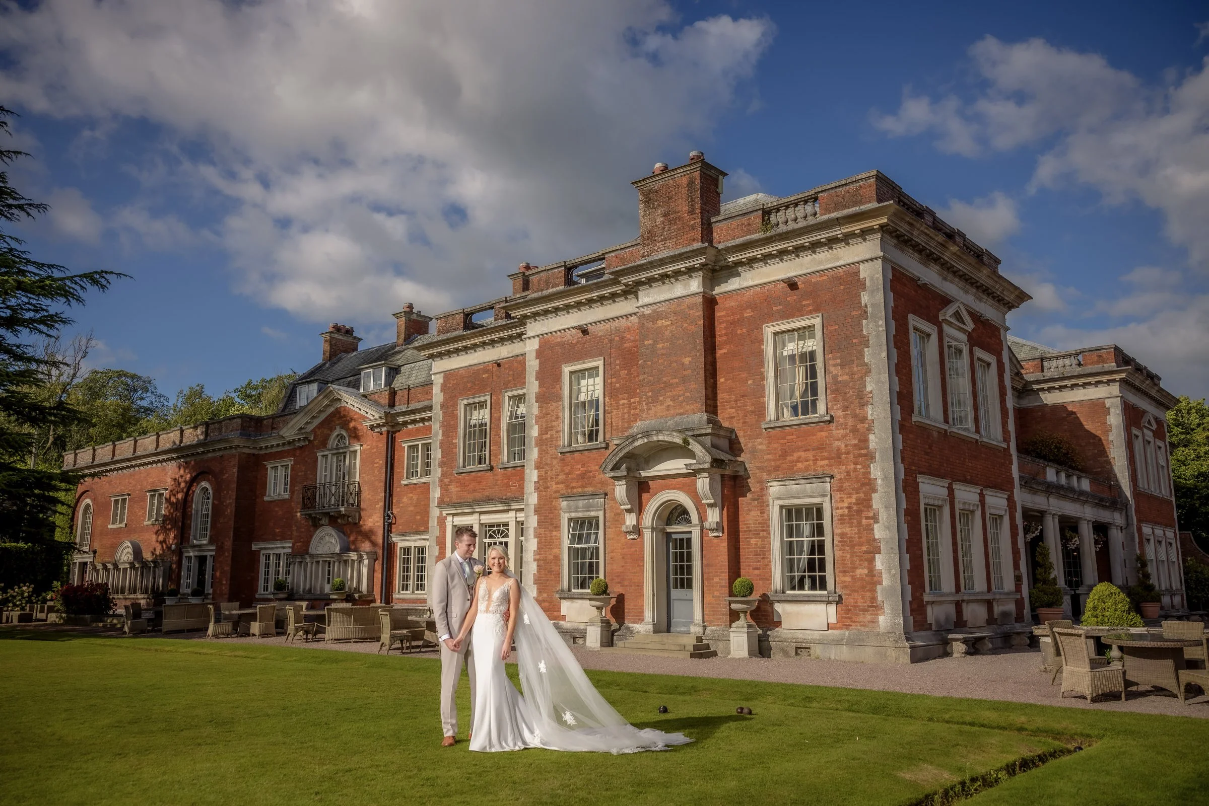 A bride and groom stand on a lawn in front of a large, historic brick mansion under a partly cloudy sky.