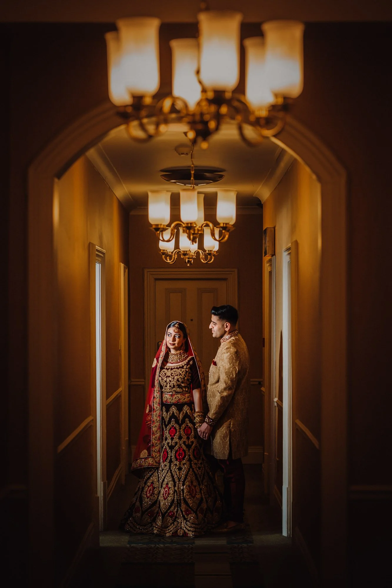 A bride and groom dressed in traditional Indian wedding attire standing hand in hand in a dimly lit corridor with ceiling lights.