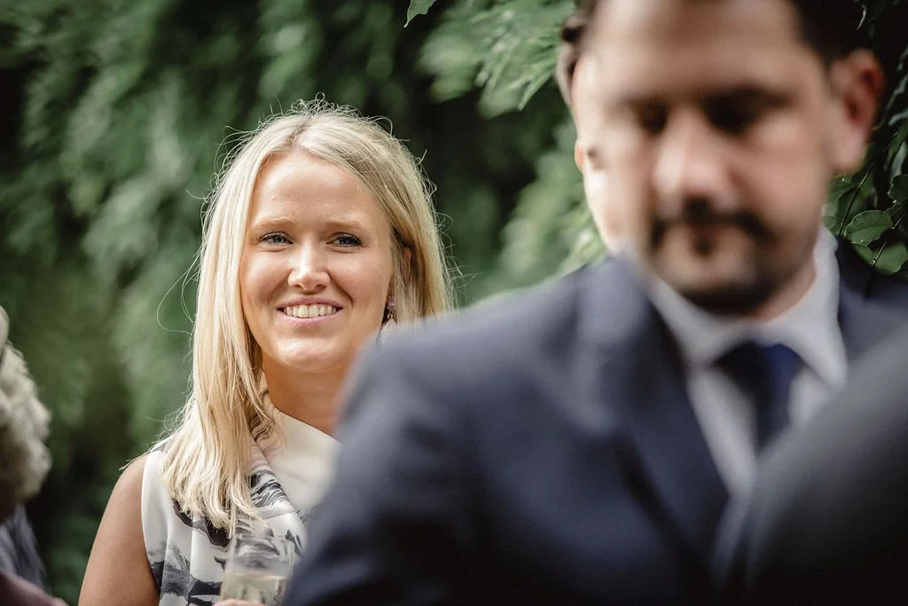 Woman smiling in a garden, with a man in a suit blurred in the foreground.